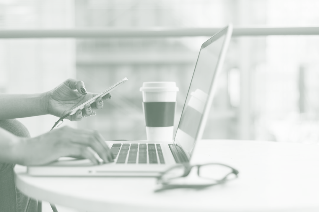 Person using a laptop while looking at smartphone with cup of coffee in the background